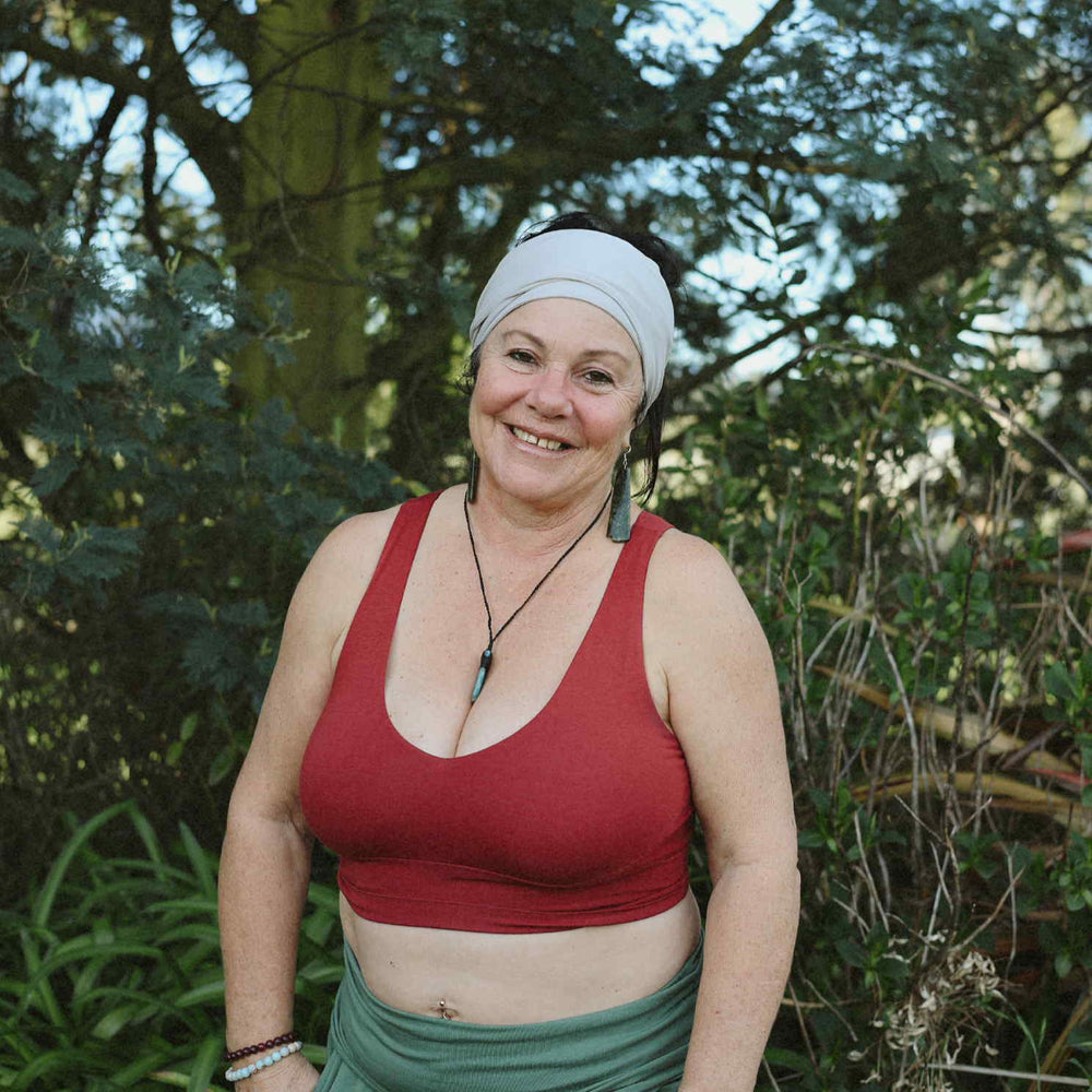 Woman wearing a red top and green pants standing in a natural setting with trees and plants.